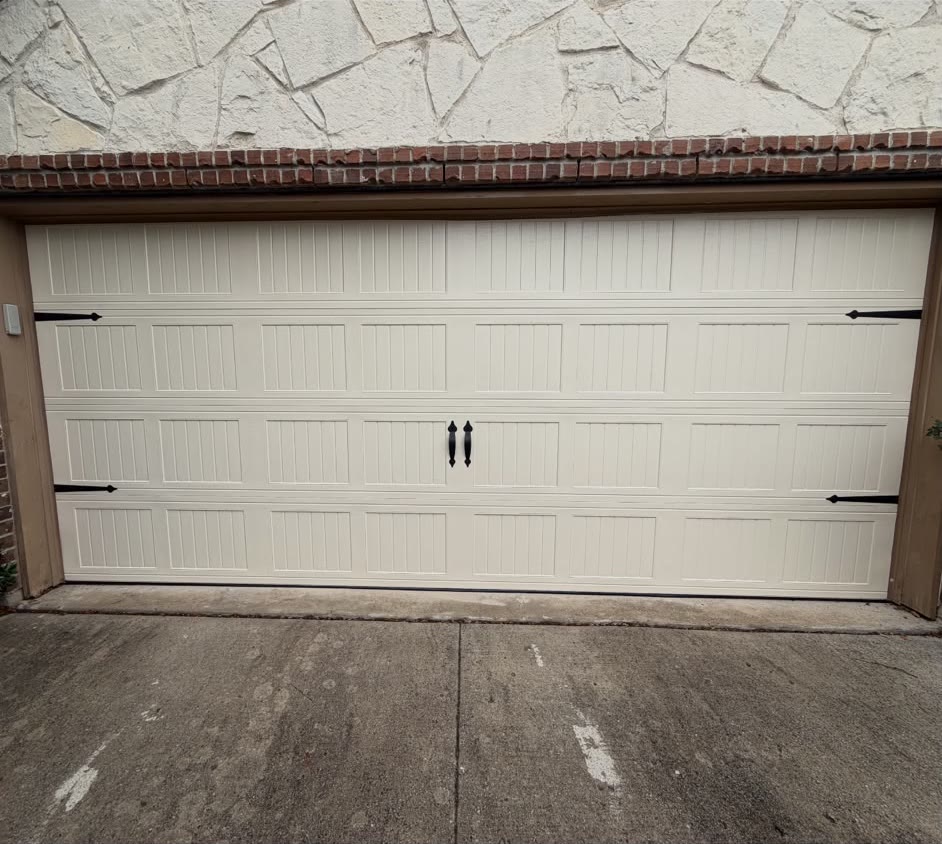 Cream carriage-house style double garage door with decorative black strap hinges and matching handles, below a white stone and red brick wall.