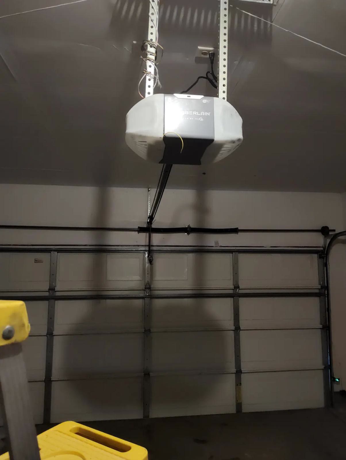 White Chamberlain myQ Wi-Fi garage door opener mounted on the ceiling of a residential garage, its light on and the rail and door arm running down to the closed sectional door below.