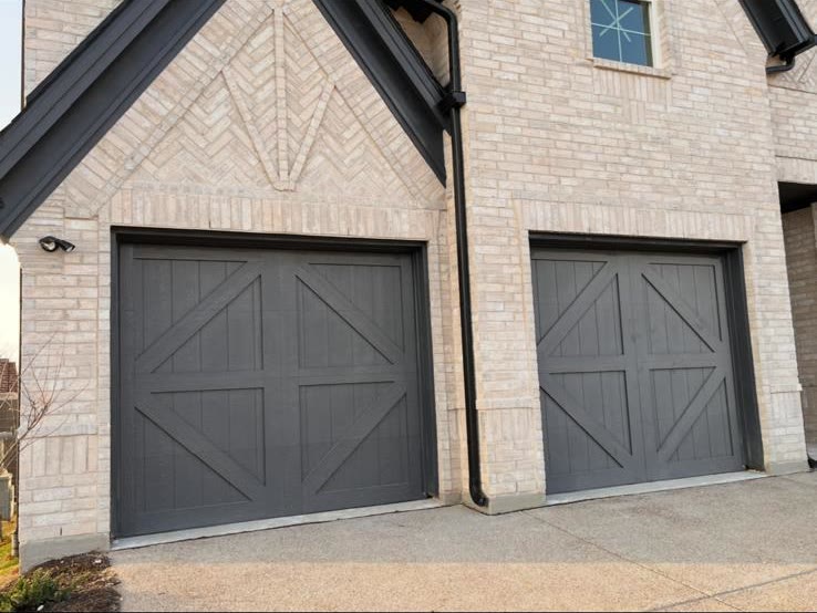 Two charcoal carriage-house style garage doors with cross-buck bracing, set into the light brick facade of a two-storey home with a steep gabled porch.