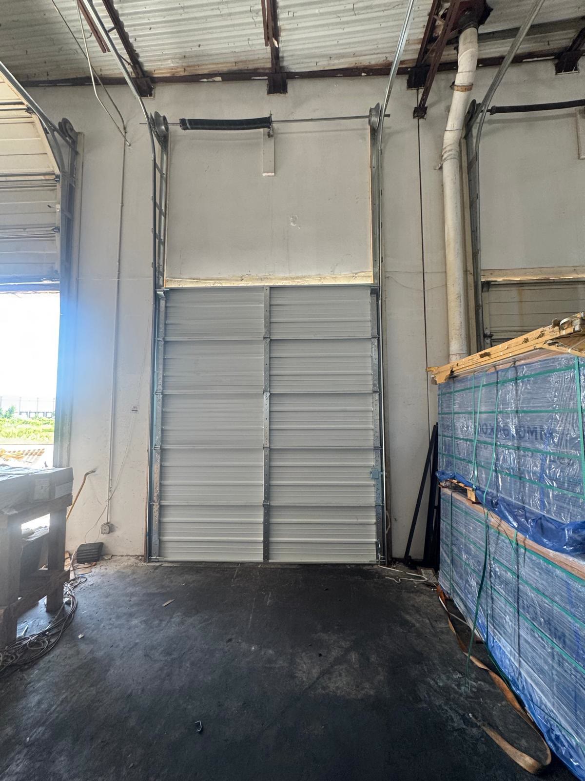 Tall commercial sectional door part-way through installation in a warehouse bay, the lower sections hung and the torsion spring and lift cables in place above, with pallets of wrapped material stacked alongside.