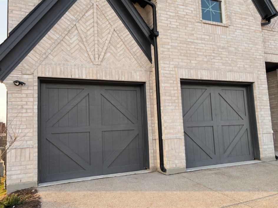 Two charcoal carriage-house style garage doors with cross-buck detailing, set into the light brick facade of a two-storey home.