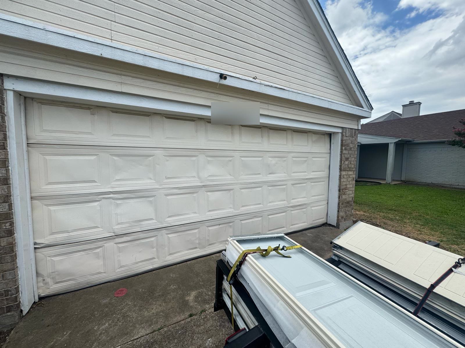 White double garage door with several creased and dented sections across its middle panels, with flat replacement panels strapped to a trailer on the driveway alongside.