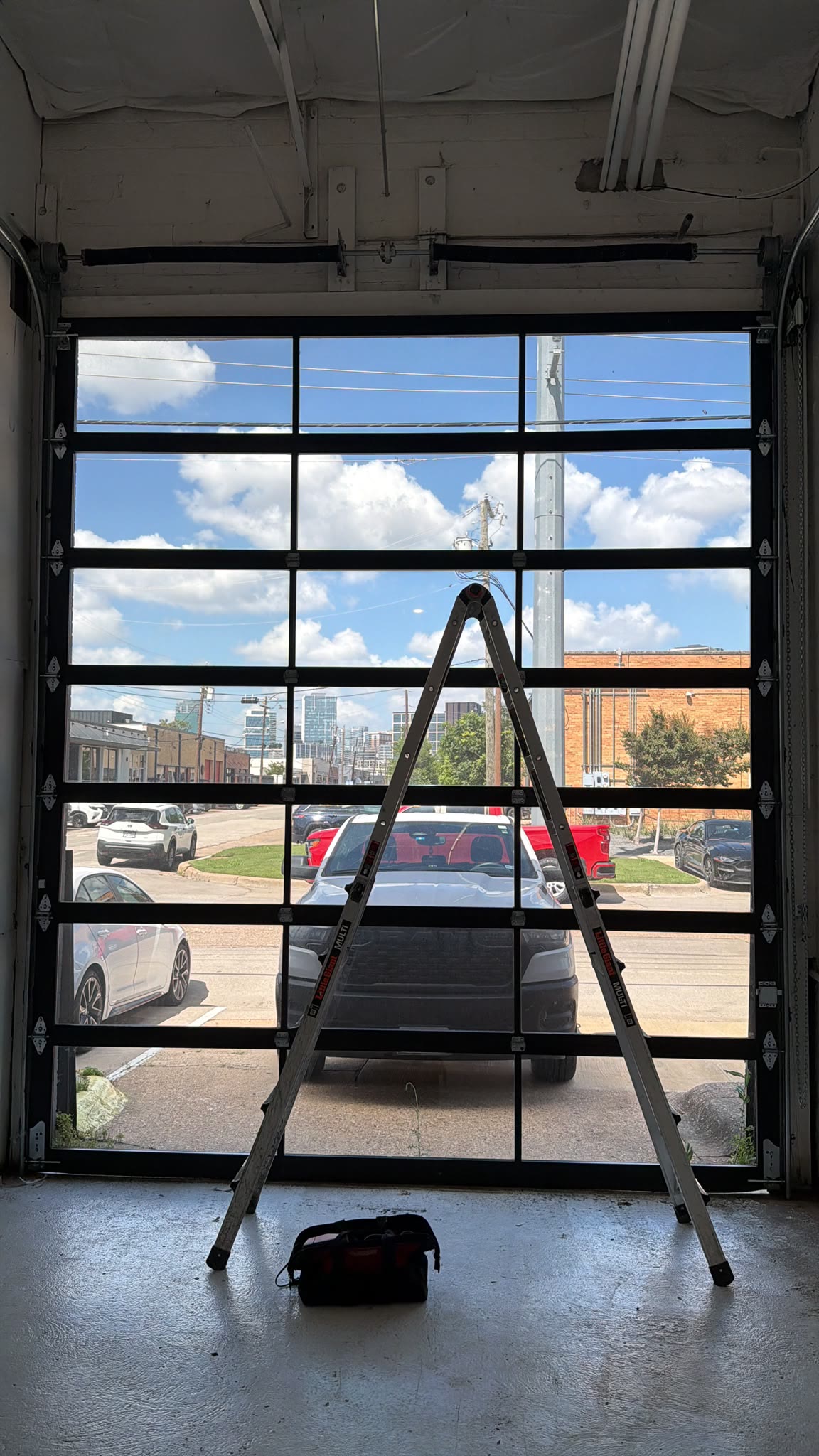 Full-view aluminium and glass commercial garage door installed in a storefront unit, with a multi-position ladder and a tool bag on the polished concrete floor and the Dallas skyline visible beyond.
