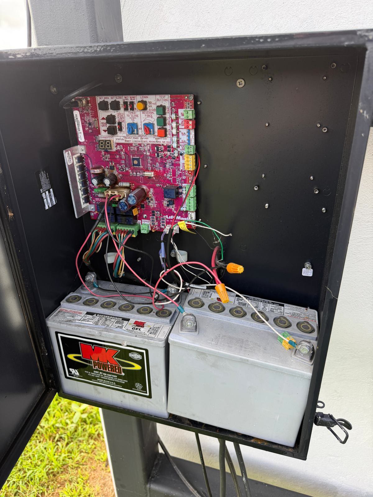 LiftMaster gate operator cabinet with the door swung open, showing the pink logic board with its open, close and reversal-force controls wired down through a loom to two sealed gel backup batteries.