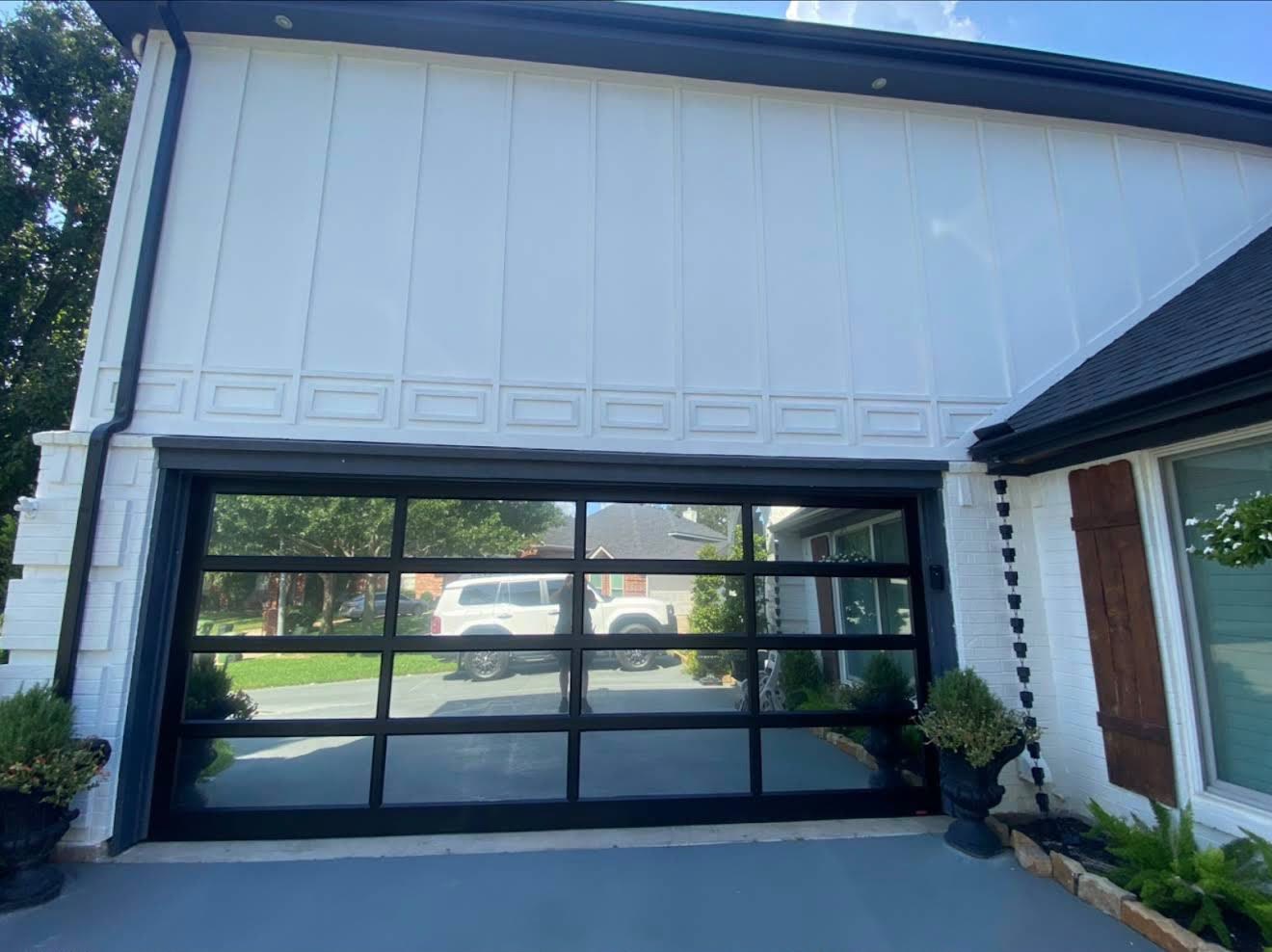 Modern full-view garage door with a black aluminium frame and clear glass panels on a white board-and-batten home, reflecting the street and trees opposite.