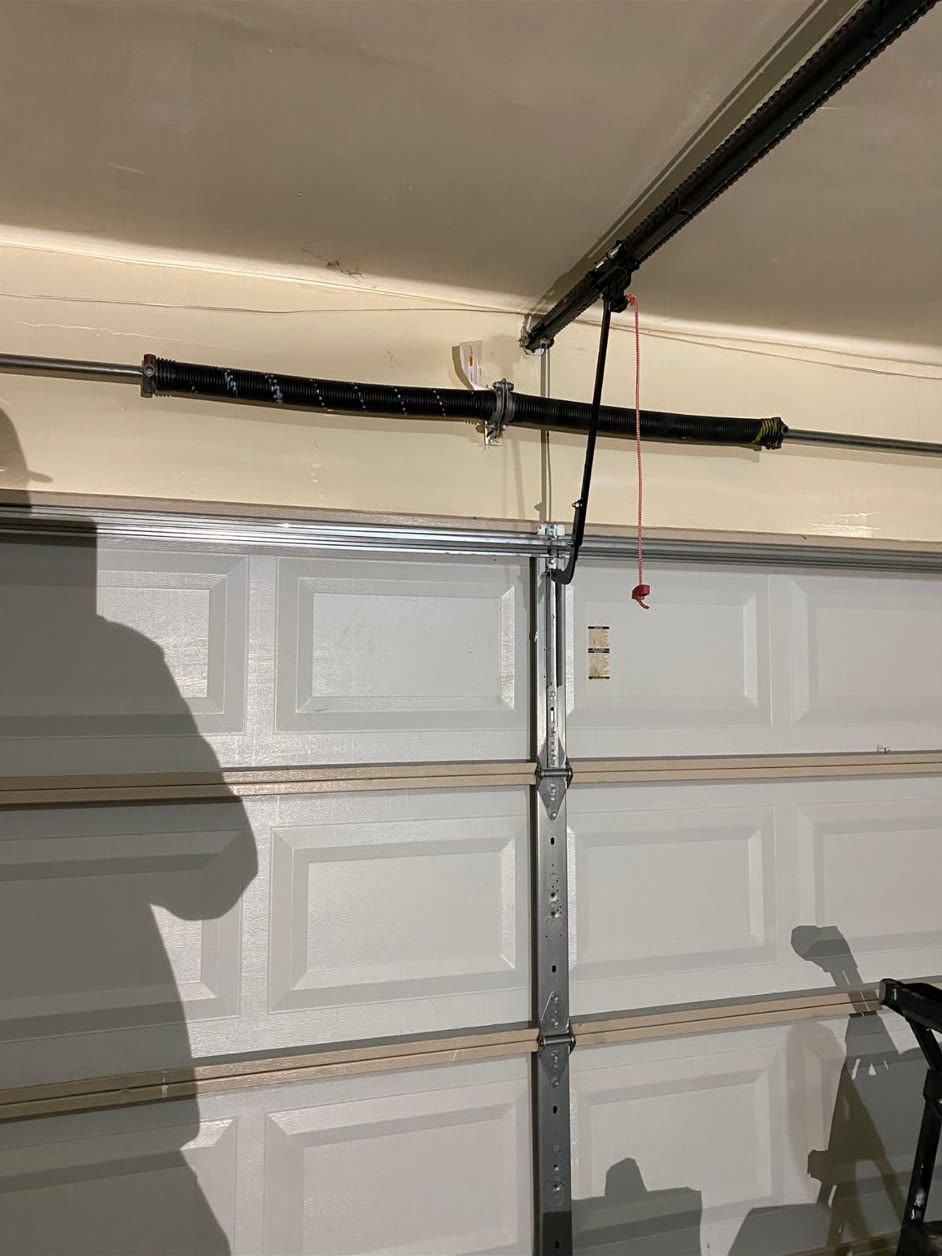 Replacement torsion spring and shaft mounted on the header above a white sectional garage door, with the red emergency release cord hanging from the opener arm.