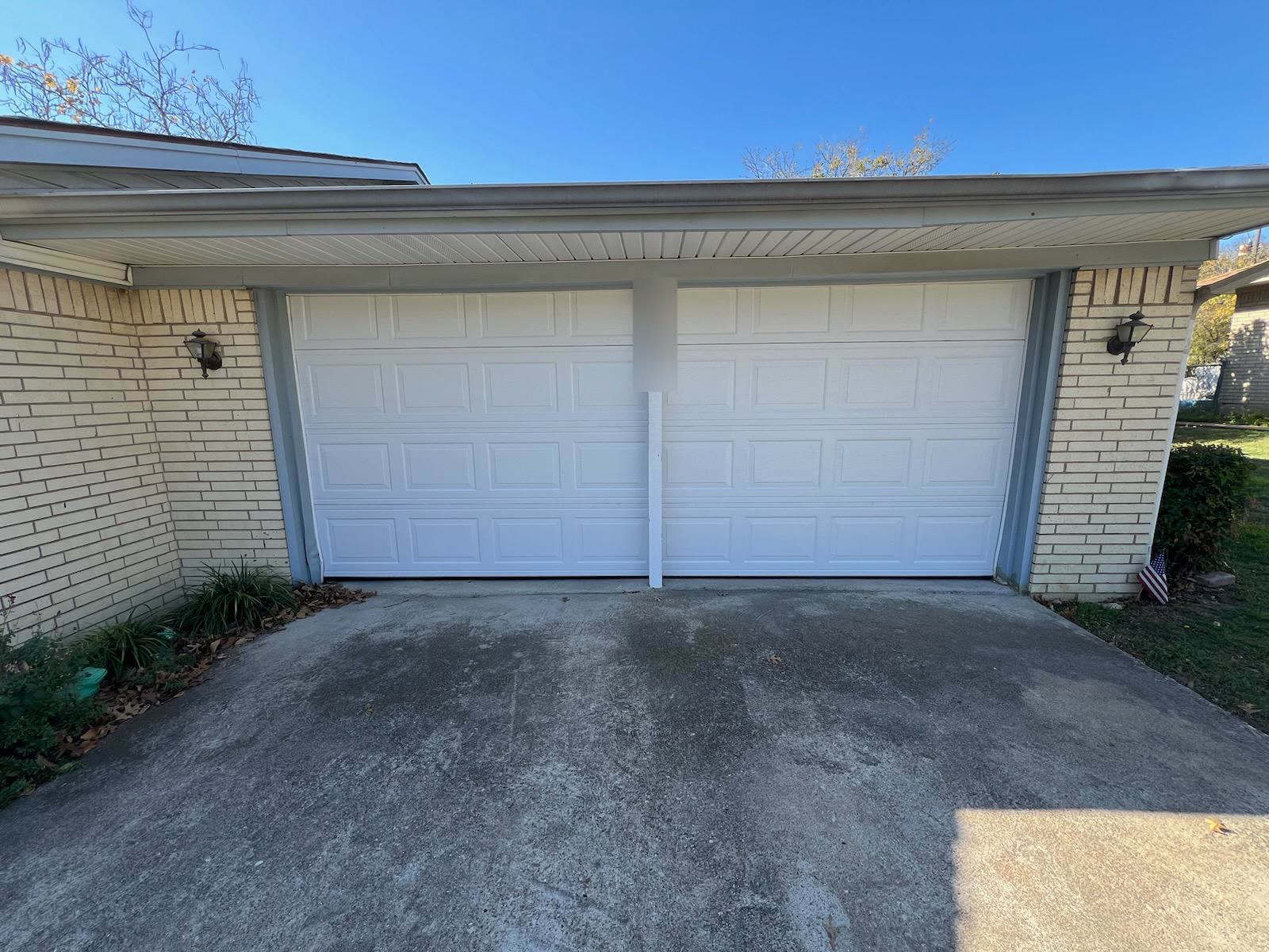 The same garage opening after repair, fitted with a new white raised-panel double door hanging square in its track, brick pillars either side.