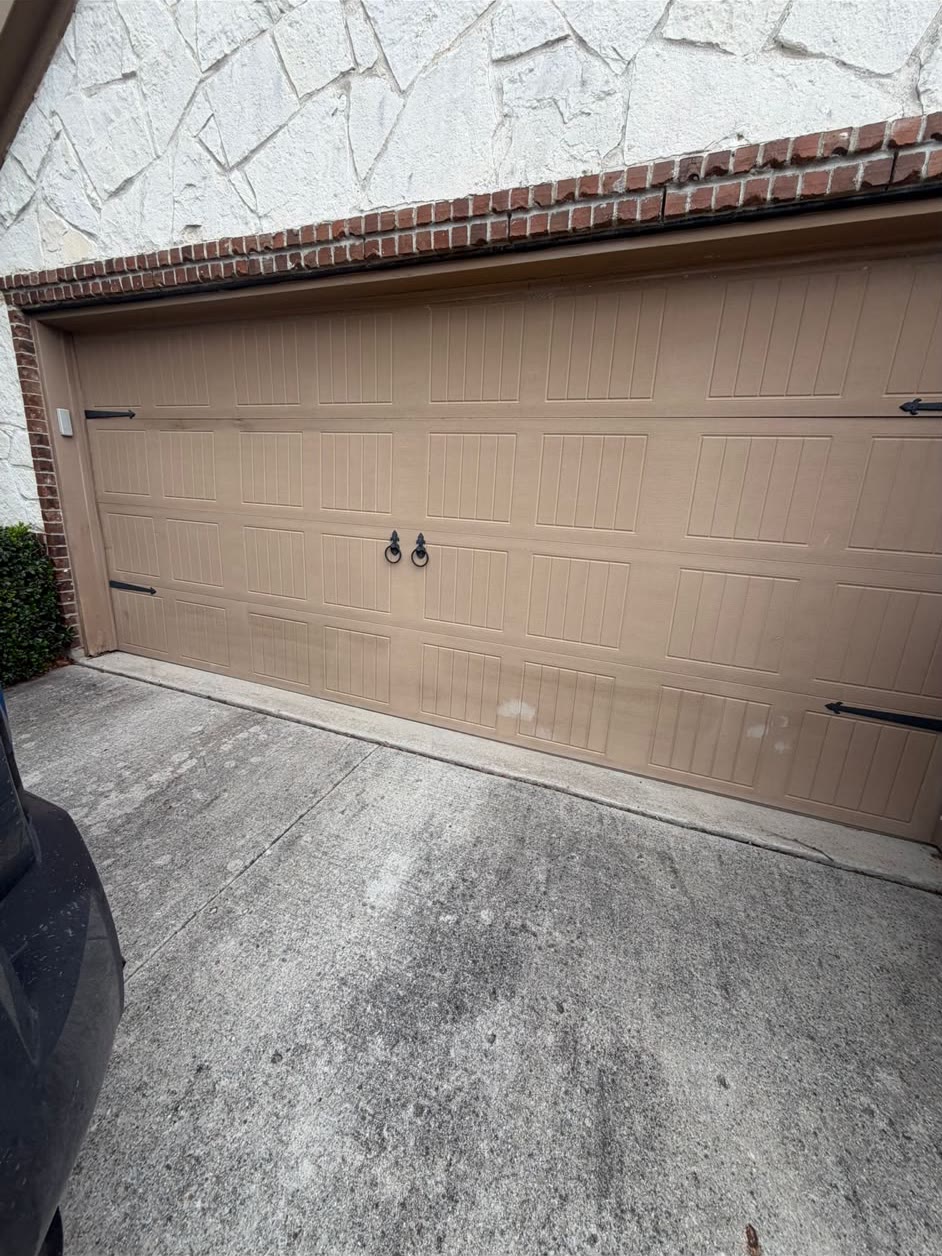Tan carriage-house style double garage door with black ring pulls and strap hinges, beneath a white stone wall trimmed with red brick.