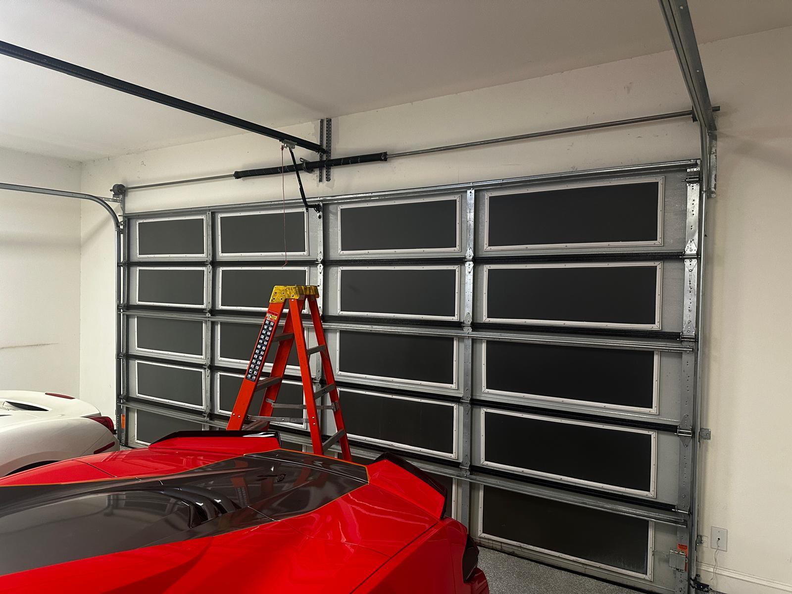 Full-view aluminium garage door with dark tinted panels seen from inside a residential garage, a red sports car and a white car parked in front of it and a step ladder standing against the door.