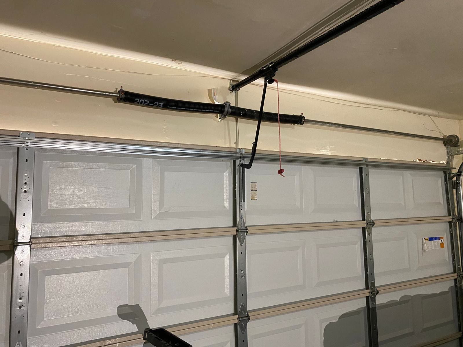 Torsion spring stencilled with its size code on the shaft above a grey raised-panel door, with the opener rail, the black curved door arm and the red emergency release cord in the same frame.