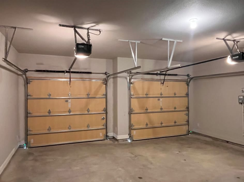 Empty two-car garage photographed from inside, with two newly installed insulated sectional doors side by side and two ceiling openers wired and mounted overhead.
