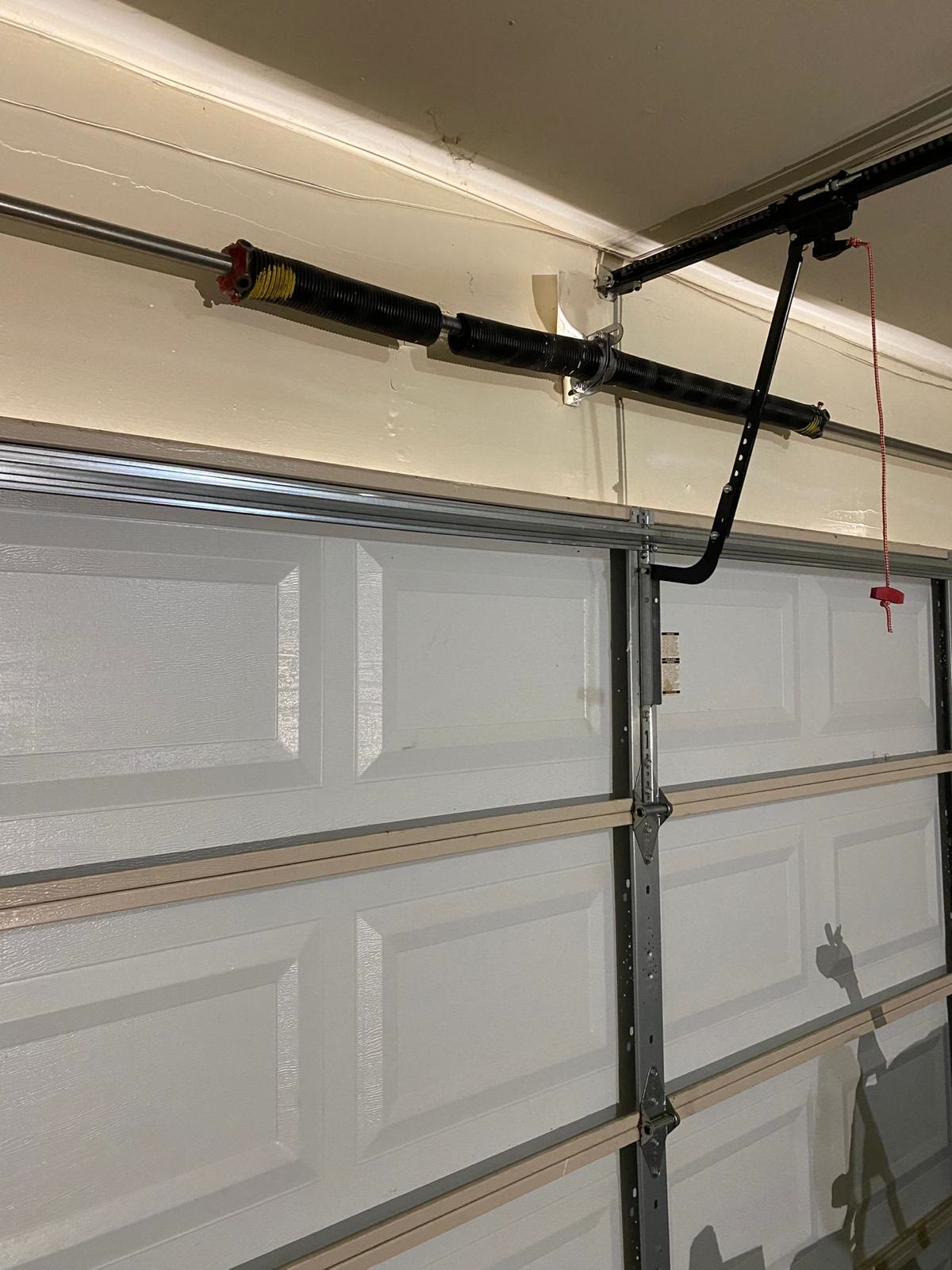 Pair of torsion springs with colour-coded winding cones on the shaft above a grey raised-panel door, alongside the opener rail, the curved door arm bolted to the top section and the red release cord.