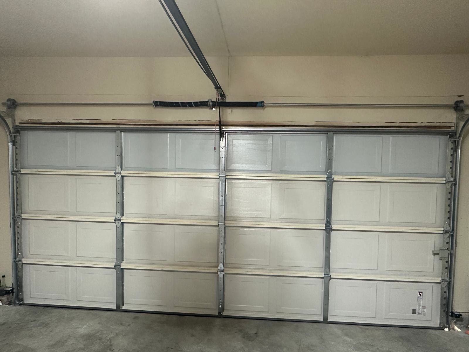 White raised-panel double garage door closed from the inside, with the torsion spring, shaft and end bearings mounted across the bare timber header above it and the opener rail coming in from the ceiling.