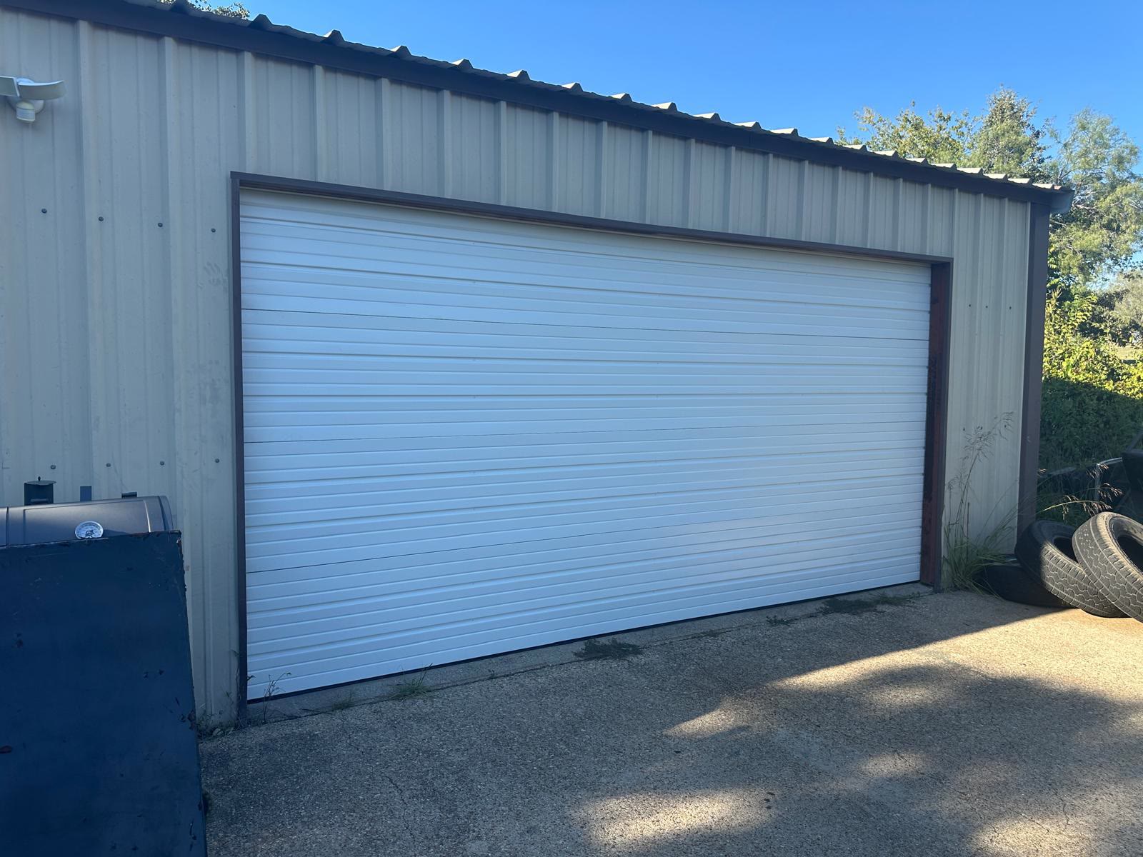 Wide white ribbed sectional door closed in the end wall of a cream steel farm building, photographed from the concrete apron under a clear blue sky.